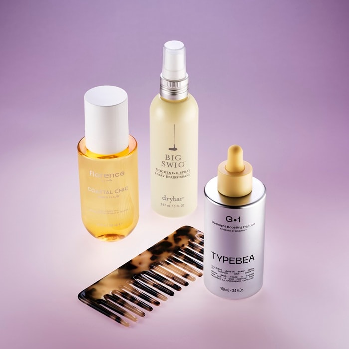 Colecție de produse pentru păr, incluzând uleiul de păr Florence într-o sticlă galbenă, spray-ul pentru volum Drybar într-o sticlă albă, serul cu peptide Typebea într-o sticlă argintie, alături de un pieptene, toate așezate pe o suprafață mov.