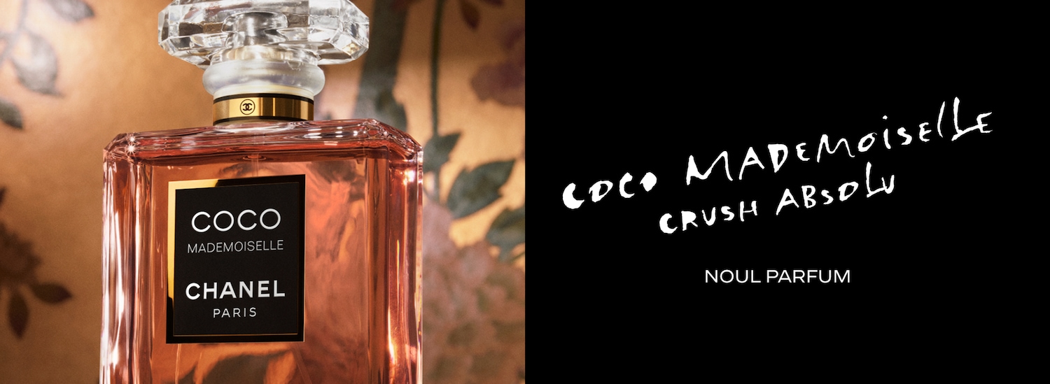 Sticla de parfum Coco Mademoiselle Crush Absolu de la Chanel, cu lichid de culoare piersică, are un capac transparent, fațetat, și o etichetă neagră cu text alb, așezată pe un fundal maro cu motive florale.