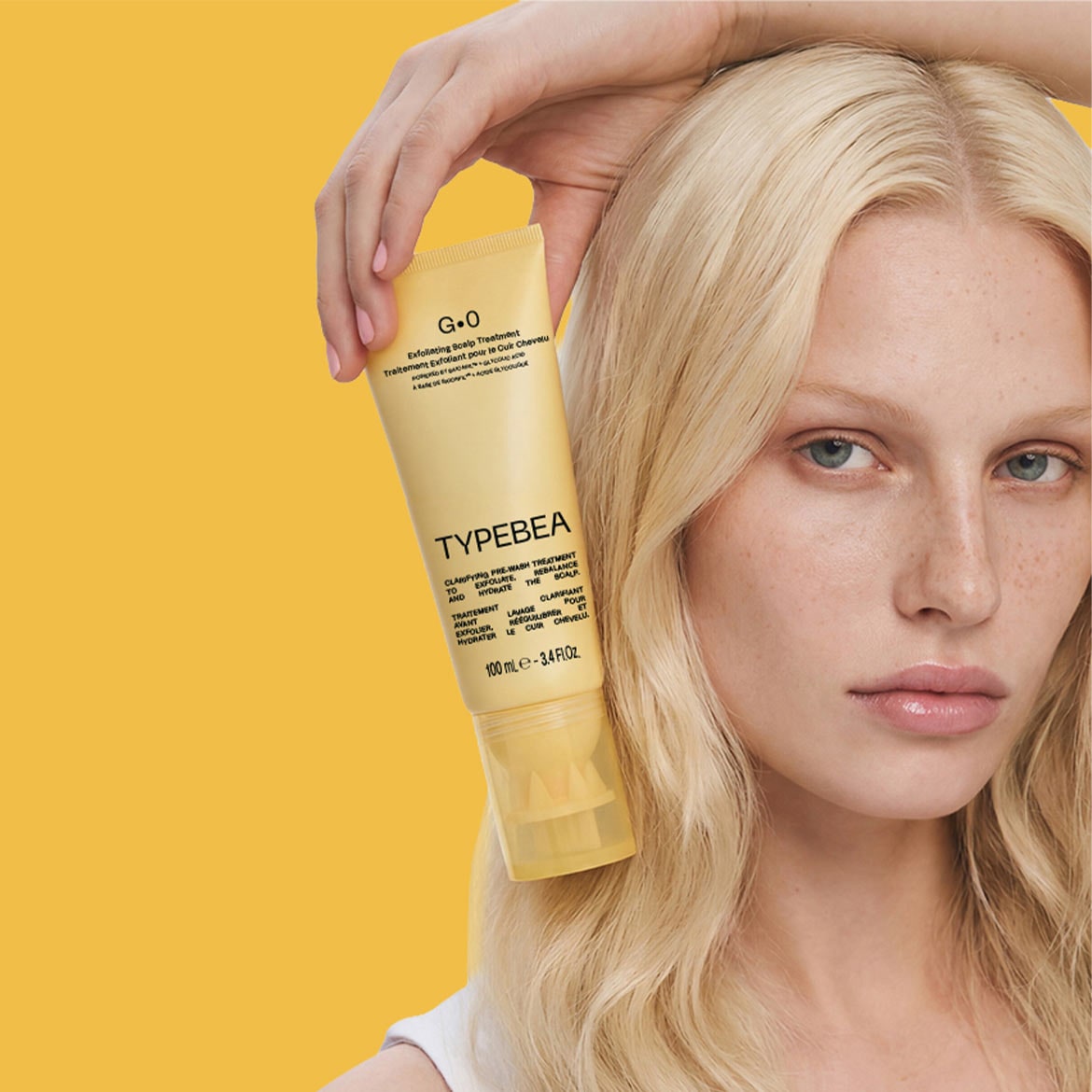 O femeie cu păr blond, cu ochi albaștri, ține în mână un tub galben de tratament exfoliant pentru scalp G.0 Typebea, cu text negru, pe un fundal galben.