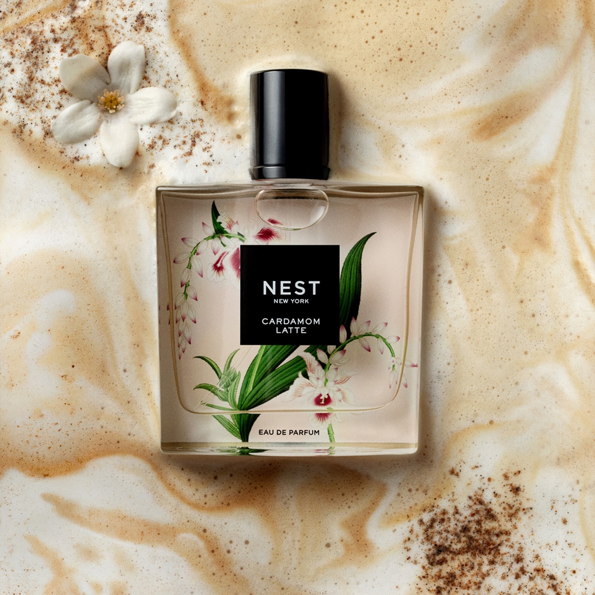 Descoperă parfumul Nest New York Cardamom Latte Eau de Parfum, o sticlă elegantă cu un design floral delicat, așezată pe o suprafață cremoasă, cu o floare albă și pudră de scorțișoară, evocând o aromă caldă și sofisticată.