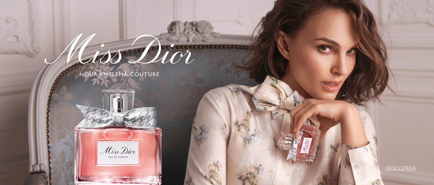 Miss Dior Noua Emblemă Couture, un parfum roz pal, cu o fundă argintie, așezat pe un scaun elegant, alături de o femeie cu păr șaten, care ține în mână o versiune miniaturală a parfumului.
