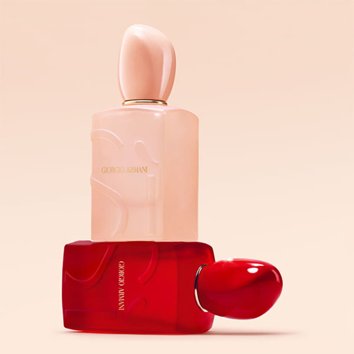 Două flacoane de parfum Armani, unul roz pal și unul roșu intens, așezate artistic pe un fundal nude.