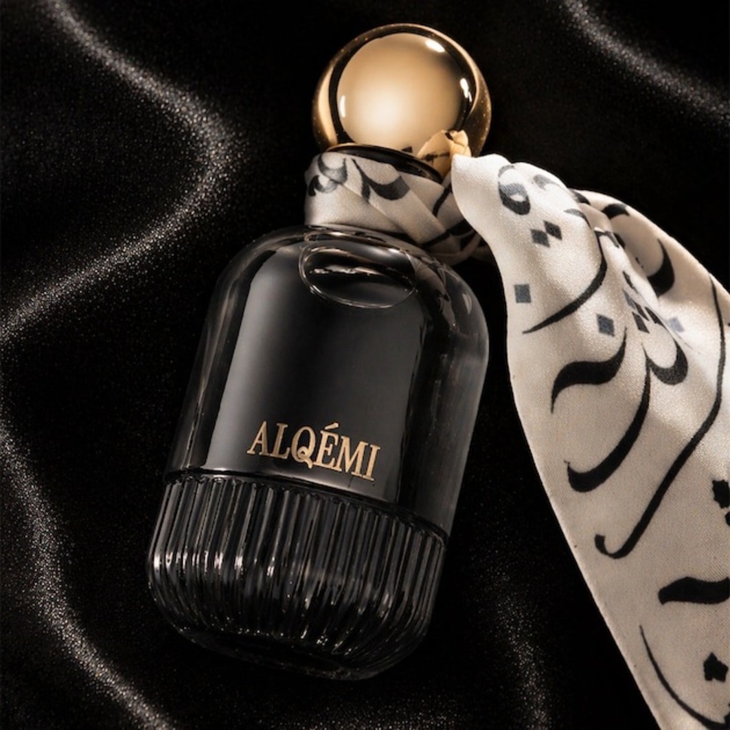 Descoperiți parfumul ALQÉMI într-o sticlă elegantă, neagră, cu un capac auriu și o eșarfă albă cu motive arabe, așezată pe un fundal de satin negru.