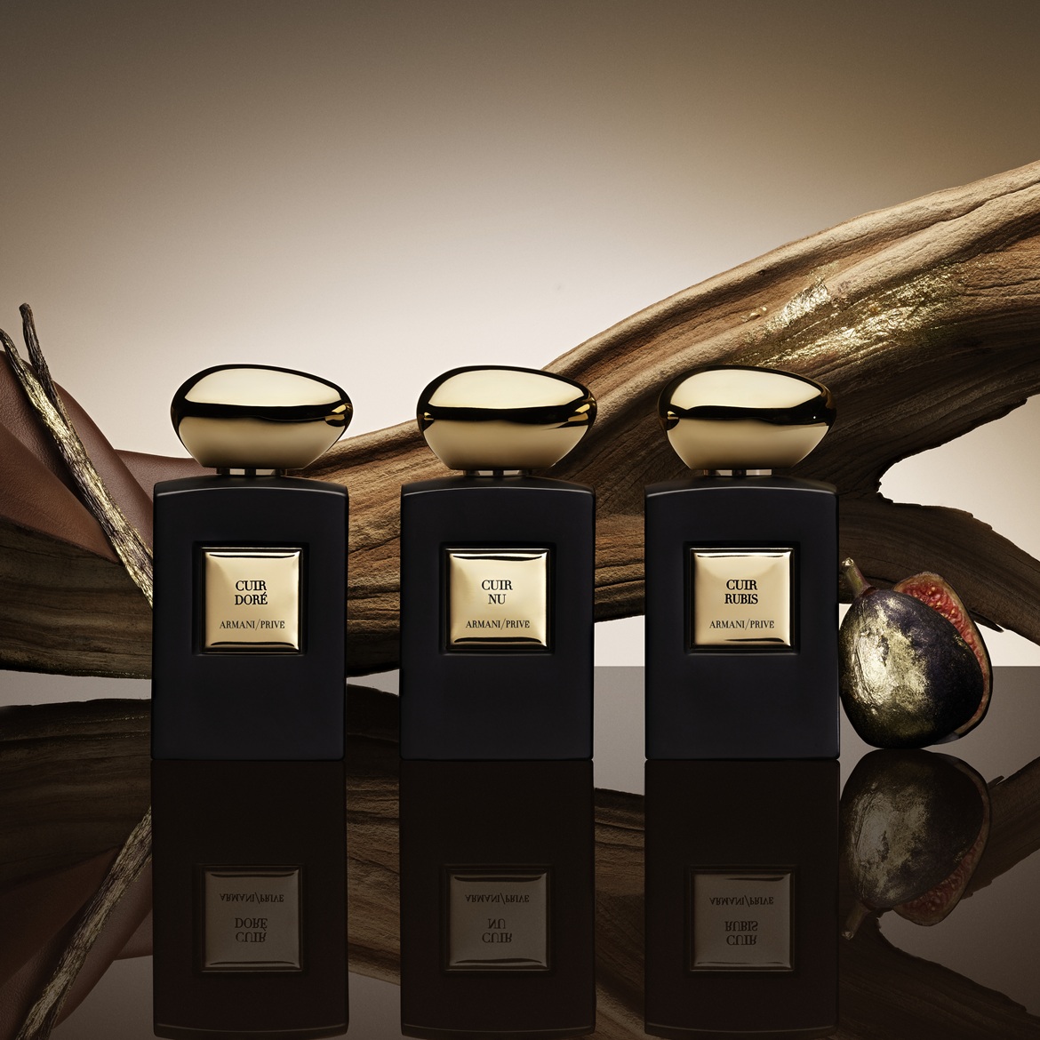 Trei sticle de parfum Armani/Privé, negre, cu etichete aurii și capace aurii, rotunde, sunt așezate pe o suprafață reflectorizantă, cu un fundal maro și elemente decorative din lemn și fructe.