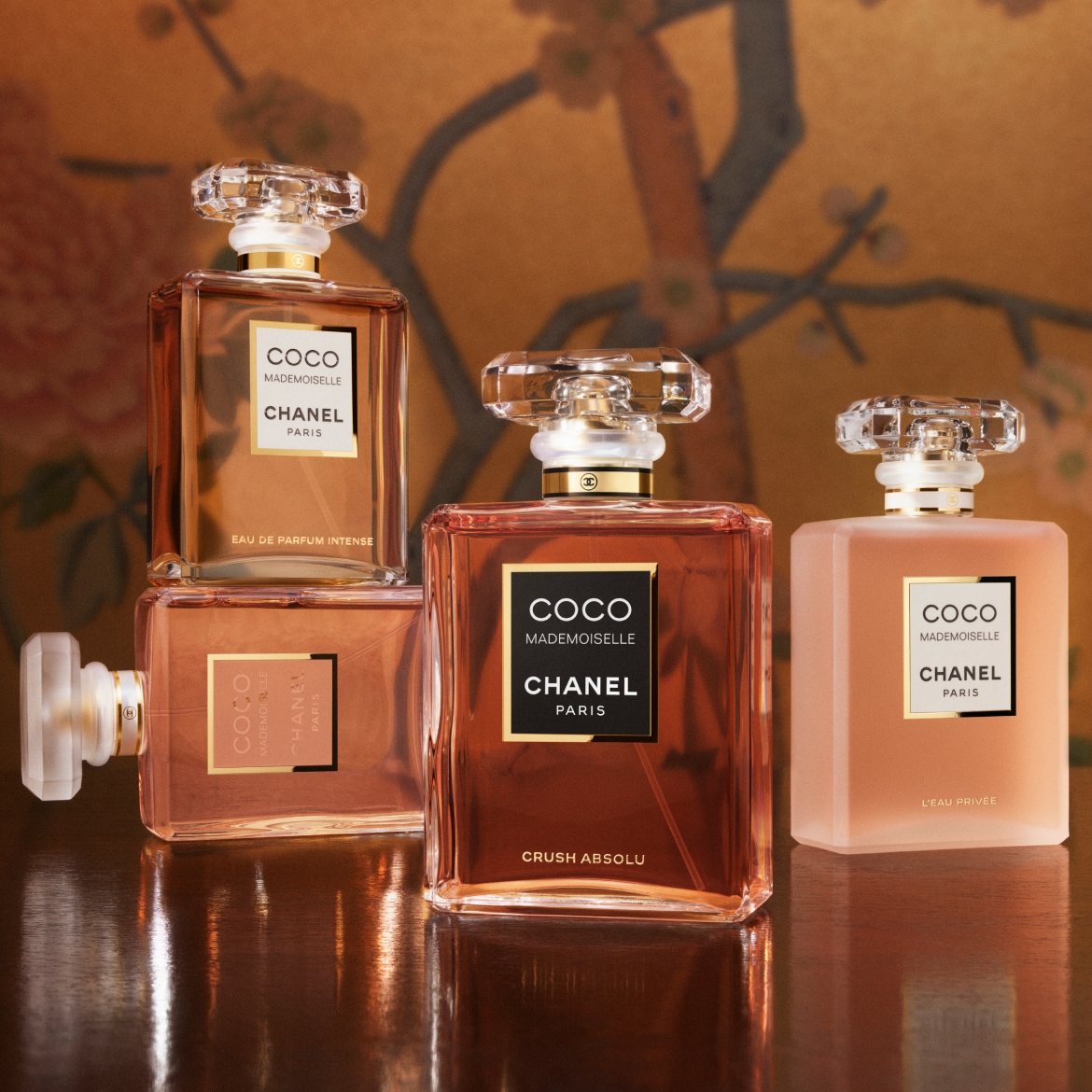 Patru sticle de parfum Coco Mademoiselle de la Chanel, cu etichete negre și aurii, sunt aranjate pe o suprafață lucioasă, cu un fundal floral estompat.
