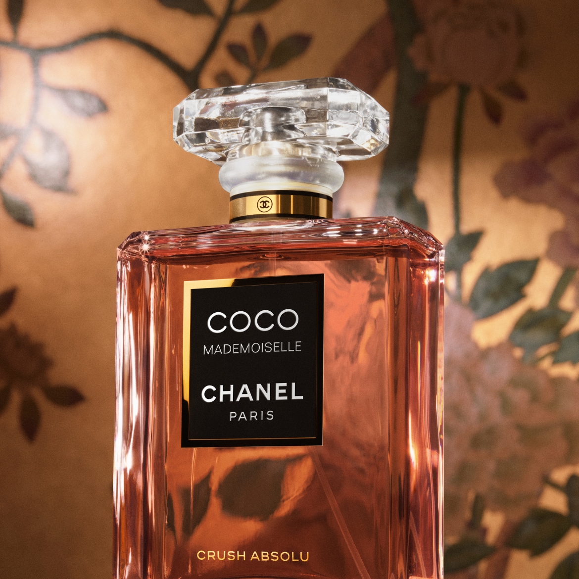 Un flacon elegant de parfum Coco Mademoiselle Chanel Paris Crush Absolu, cu lichid de culoare piersică, capac transparent fațetat și etichetă neagră cu text alb, așezat pe un fundal auriu cu motive florale.