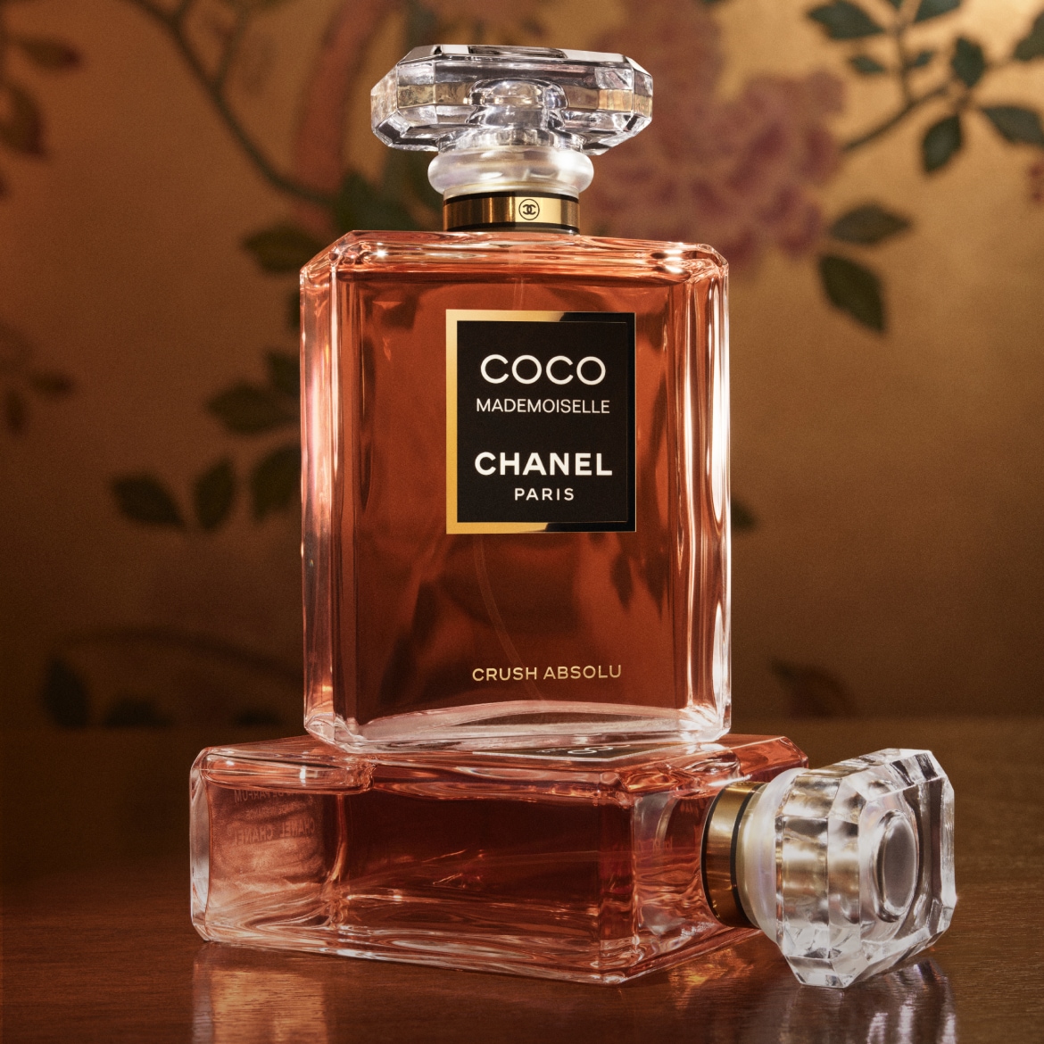 Descoperiți parfumul Coco Mademoiselle Chanel Crush Absolu, o esență elegantă într-o sticlă pătrată, cu lichid de culoarea chihlimbarului și un capac transparent, fațetat, așezată pe o masă de lemn, cu un fundal floral estompat.