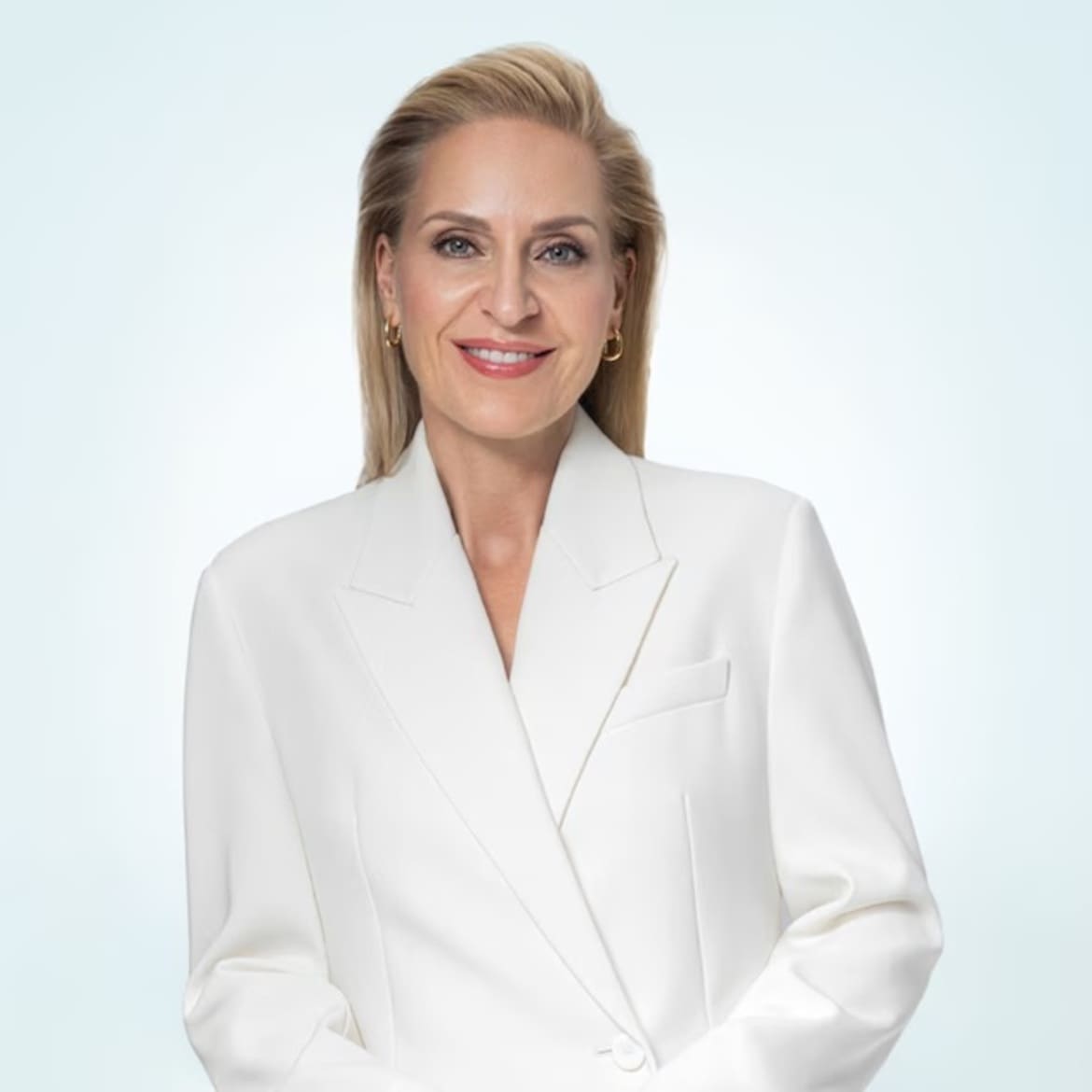 O femeie blondă, cu părul prins la spate, poartă un sacou alb și cercei aurii, zâmbind cu buze roșii.