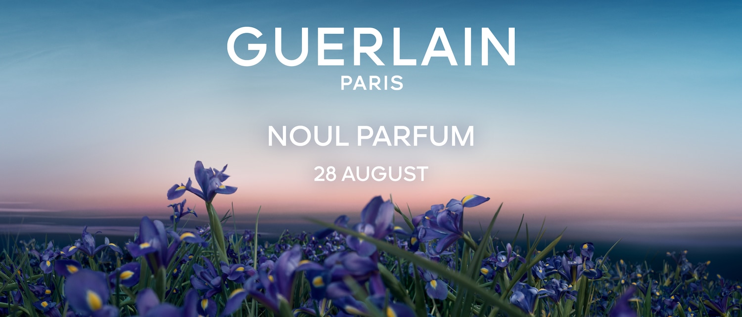 Imaginea prezintă un câmp de flori de iris violet cu centre galbene, sub un cer albastru-rozaliu, cu textul "GUERLAIN PARIS NOUL PARFUM 28 AUGUST" în alb.