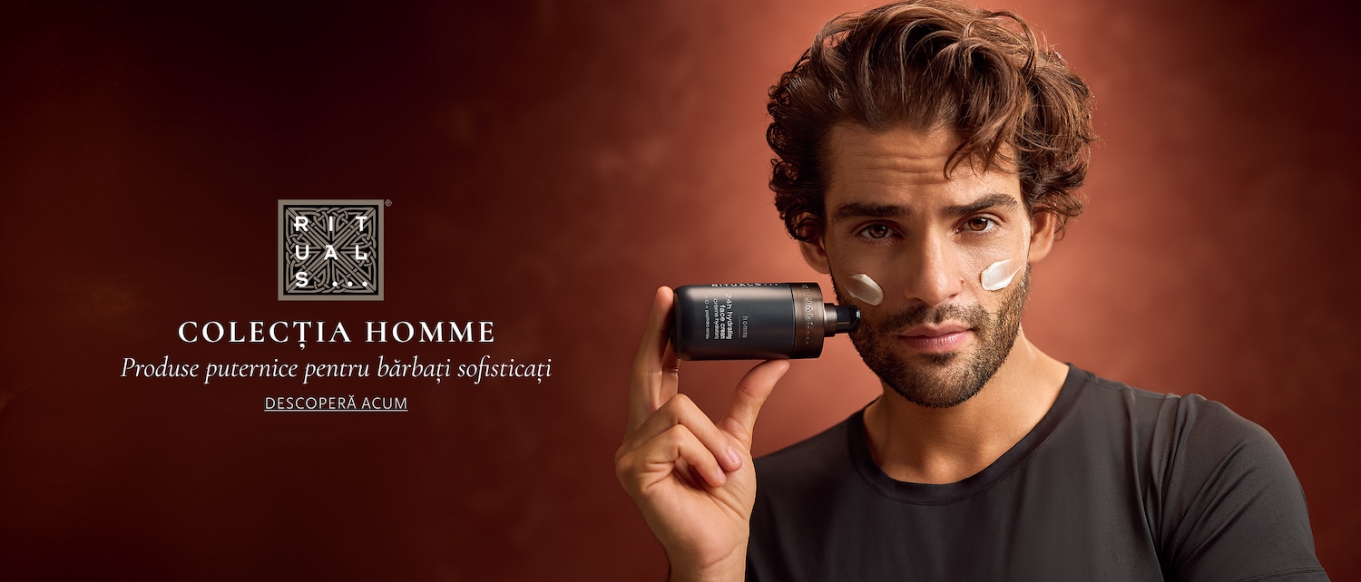 Un bărbat cu păr șaten, creț, cu barbă, poartă un tricou negru și aplică o cremă hidratantă de față Rituals Homme 24h, cu textură albă, pe ambii obraji, având pe fundal un perete maro.