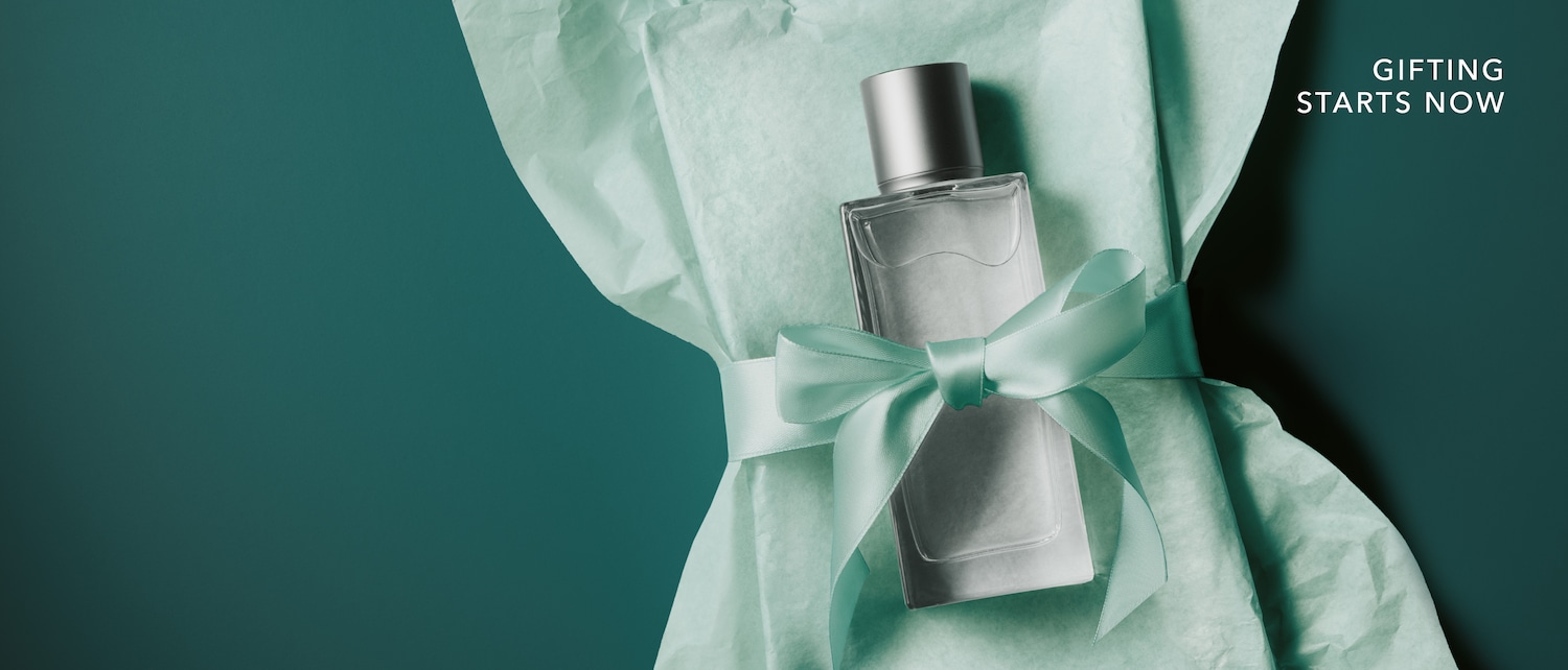 Un flacon de parfum elegant cu capac argintiu este împachetat în hârtie de mătase verde-mentă și legat cu o fundă satinată, însoțit de mesajul Gifting starts now. Cumpără acum cadoul perfect!
