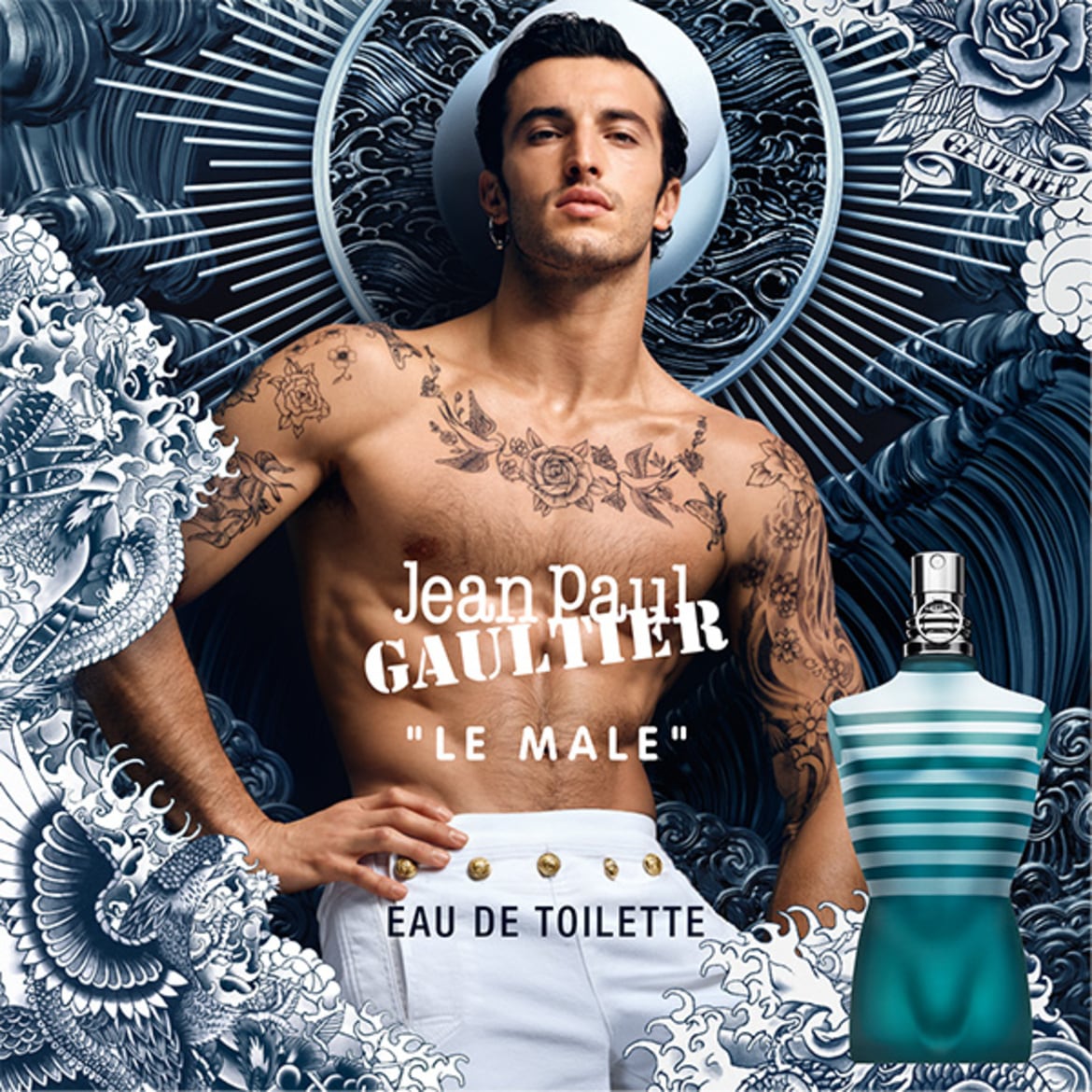 Un bărbat tatuat, cu o pălărie de marinar albă, pozează alături de sticla de parfum Jean Paul Gaultier "Le Male" Eau de Toilette, pe un fundal albastru închis cu motive florale.
