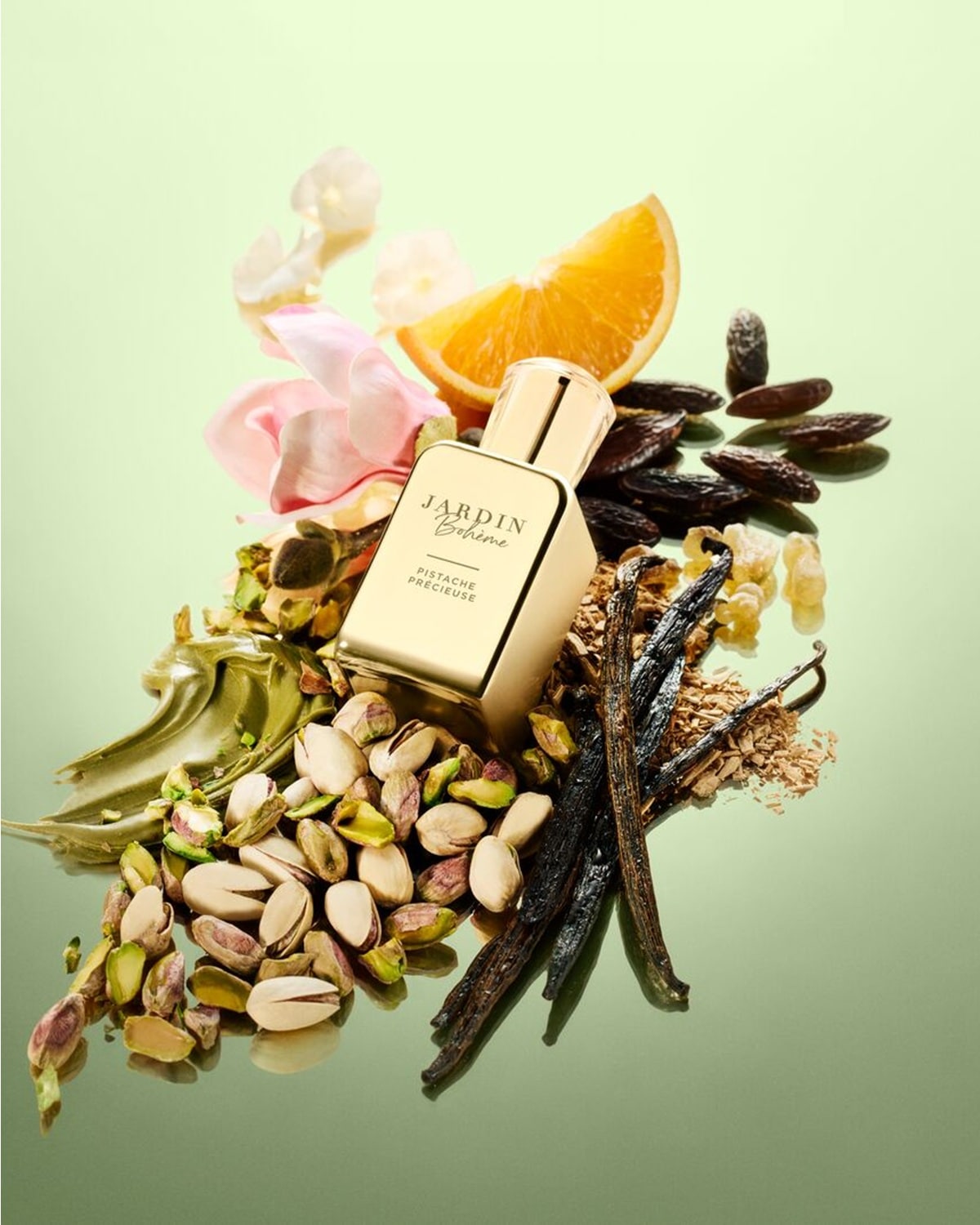 Un flacon auriu de parfum Jardin Bohème Pistache Précieuse este așezat printre fistic, păstăi de vanilie, o felie de portocală, petale de flori roz și boabe de tonka, pe un fundal verde deschis.