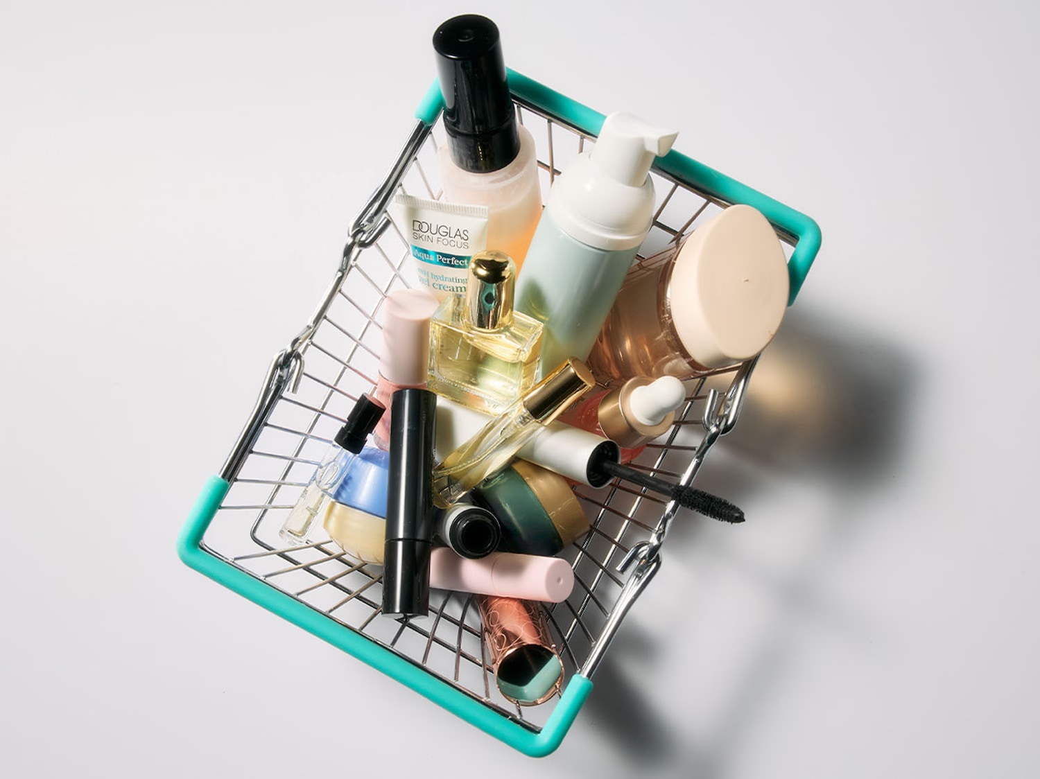 Un coș de cumpărături metalic, cu mânere turcoaz, este plin cu diverse produse cosmetice, inclusiv o cremă hidratantă Douglas Skin Focus Aqua Perfect, parfumuri, un rimel negru și alte articole de machiaj, toate așezate pe un fundal alb.