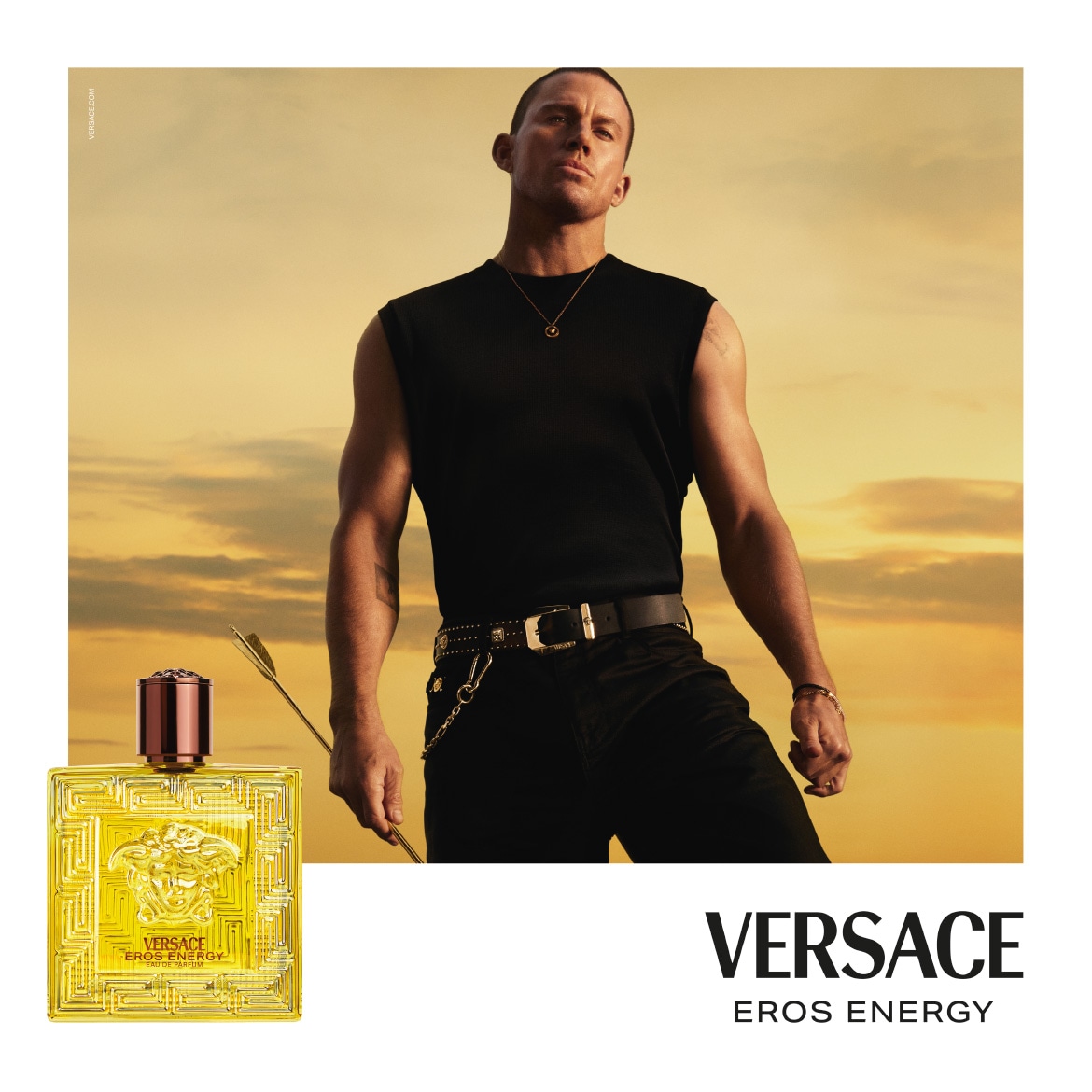 Channing Tatum poartă un maiou negru, pantaloni negri și o curea neagră cu ținte, ținând o săgeată aurie, cu un flacon de parfum Versace Eros Energy în prim-plan, pe un fundal de apus de soare.