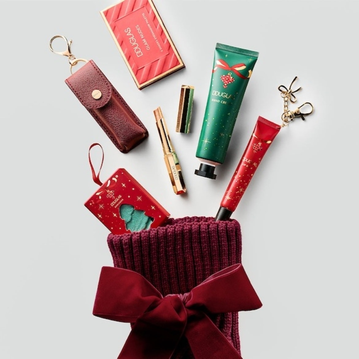 Un ciorap de Crăciun grena, decorat cu o fundă mare din catifea, conține diverse produse cosmetice festive Douglas, cum ar fi palete de farduri, rujuri aurii și creme de mâini în ambalaje verzi și roșii. Cumpără acum cadourile perfecte!