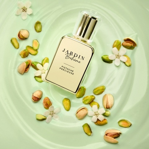 O sticlă elegantă de parfum Jardin Bohème Pistache Précieuse, cu un capac auriu și lichid de culoarea șampaniei, este așezată pe o suprafață verde deschis, înconjurată de fistic și flori albe delicate.