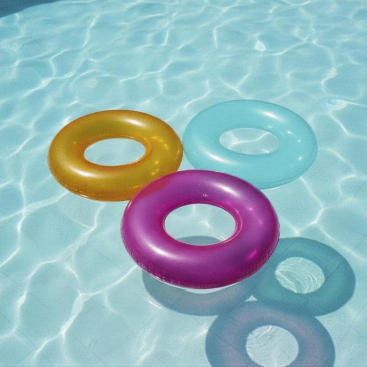 Trei inele de înot colorate, unul auriu, unul albastru deschis și unul magenta, plutesc pe suprafața apei cristaline a unei piscine, creând reflexii luminoase.