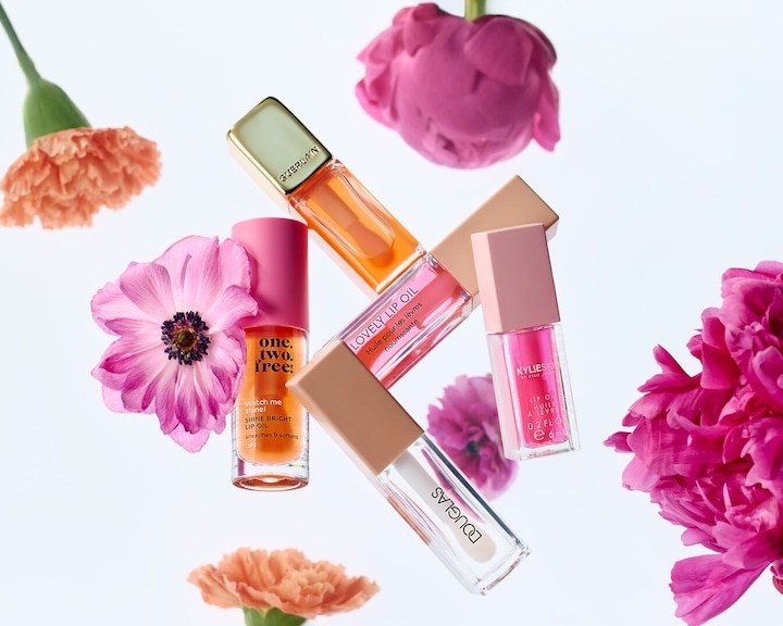 Mai multe uleiuri de buze de la Guerlain, Kylie Jenner și Douglas sunt expuse alături de flori colorate, evidențiind nuanțele lor vibrante și texturile lucioase.