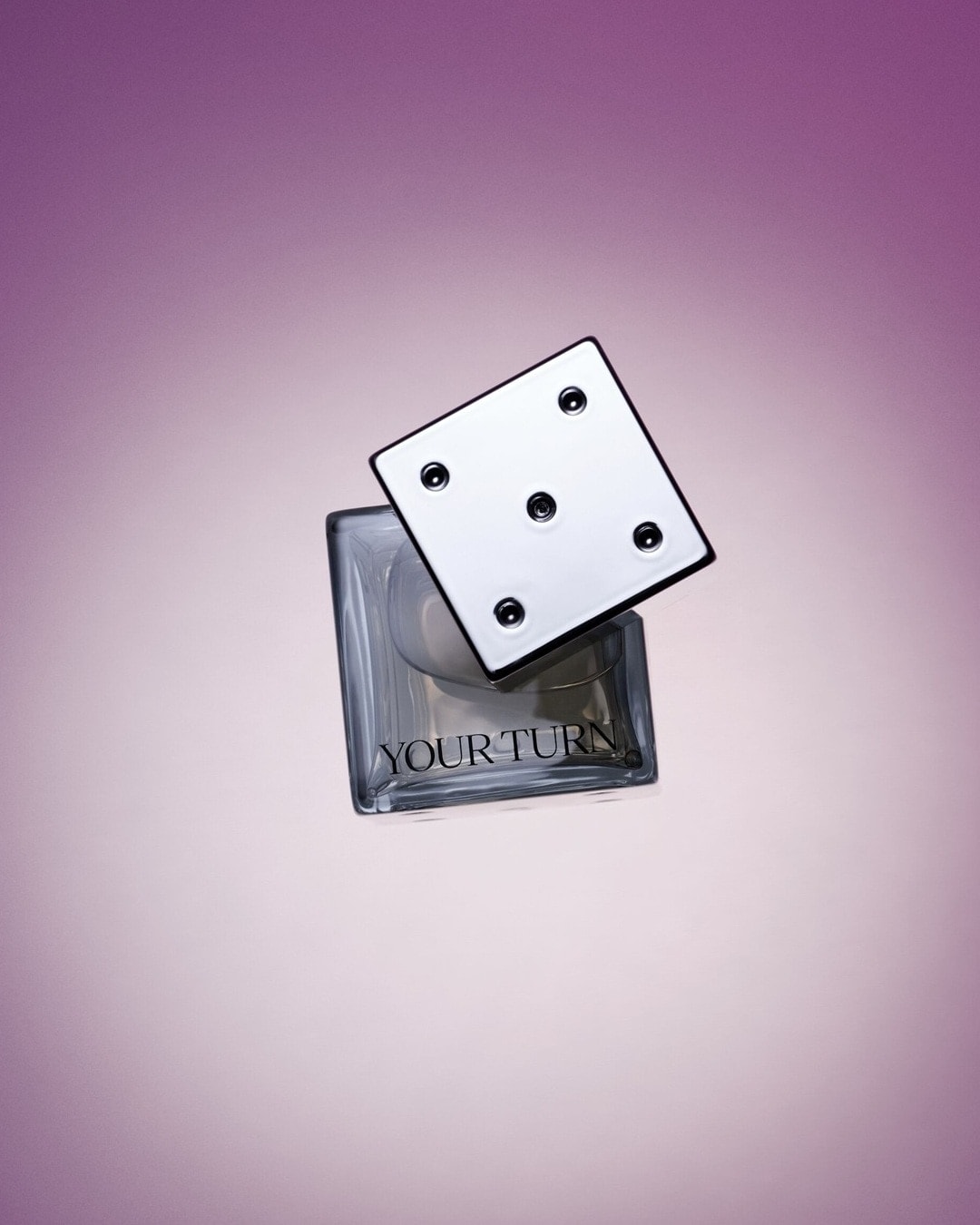 O sticlă de parfum cu un capac în formă de zar, așezată pe un fundal mov în degrade, cu eticheta „YOUR TURN".