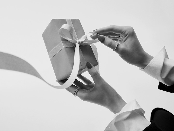 Mâinile desfac o panglică de pe o cutie cadou împachetată, invitându-te să descoperi cadoul perfect la Douglas.