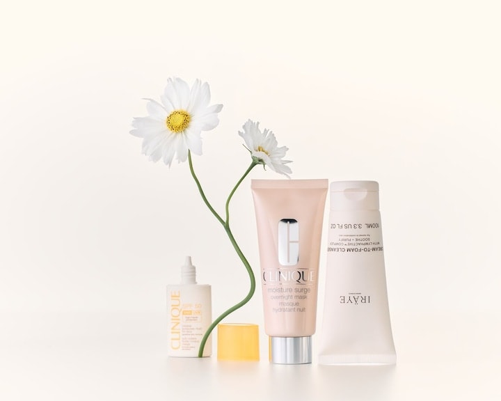 Sunt prezentate produse de îngrijire a pielii de la Clinique și IRAYE, inclusiv masca Clinique Moisture Surge într-un tub roz deschis și cleanserul cremă-spumă de la IRAYE, completate de flori albe delicate.