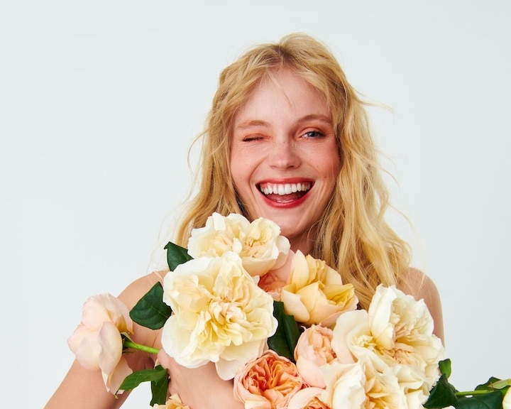 O femeie blondă face cu ochiul, ținând un buchet de trandafiri în nuanțe de galben deschis și piersică, perfect pentru îngrijirea pielii în primăvară.
