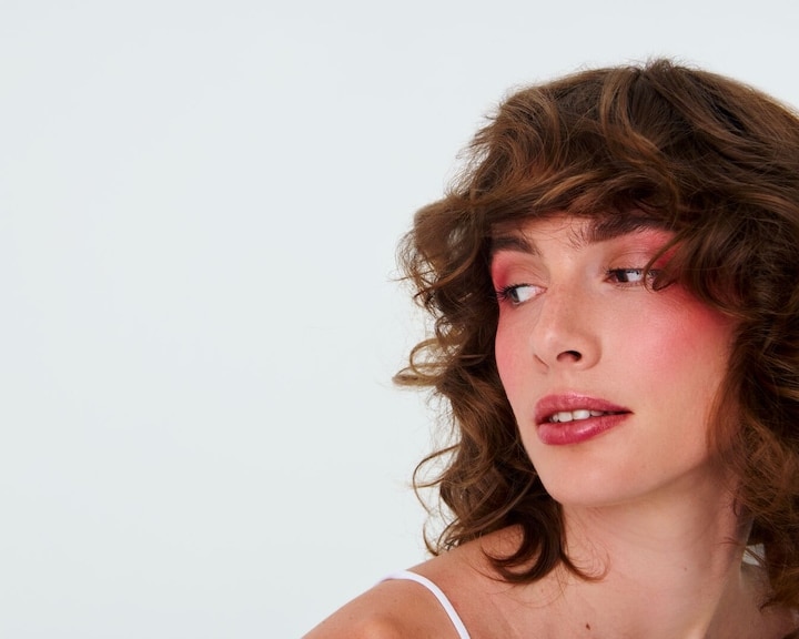 O femeie cu păr creț șaten și machiaj roz pe pleoape și obraji privește în lateral, evidențiind un look proaspăt și vibrant de primăvară.