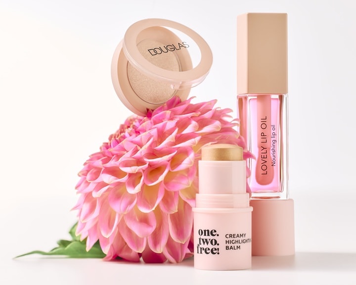 Un set de machiaj Douglas, în ambalaj roz deschis, care include o pudră strălucitoare, un balsam iluminator cremos și un ulei de buze hrănitor, toate expuse alături de o floare roz vibrantă.