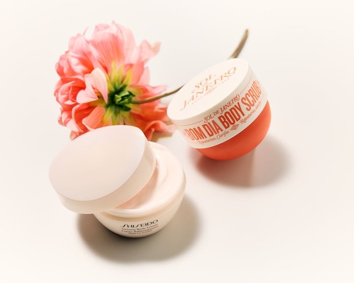 Două produse de îngrijire a pielii, inclusiv Sol de Janeiro 'Bom Dia Body Scrub' într-un recipient portocaliu, alături de o cremă Shiseido, completate de o floare roz delicată.