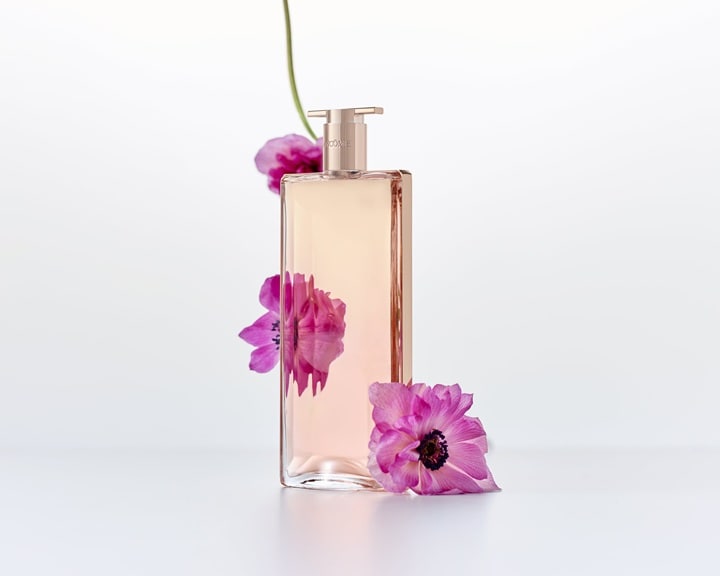 O sticlă de parfum dreptunghiulară, cu capac auriu, este împodobită cu anemone roz vibrante, creând o imagine proaspătă și elegantă, specifică unui parfum de primăvară.
