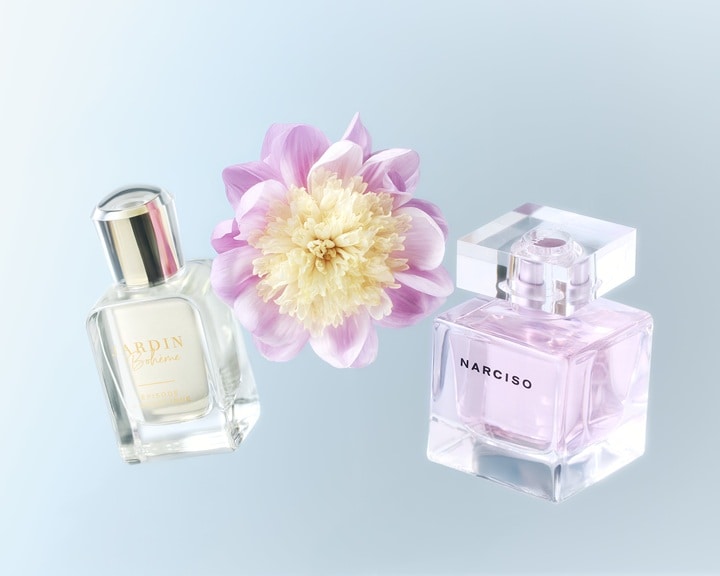 Două sticle de parfum, una transparentă cu capac argintiu și cealaltă o sticlă pătrată roz deschis, sunt expuse lângă o floare roz și albă.