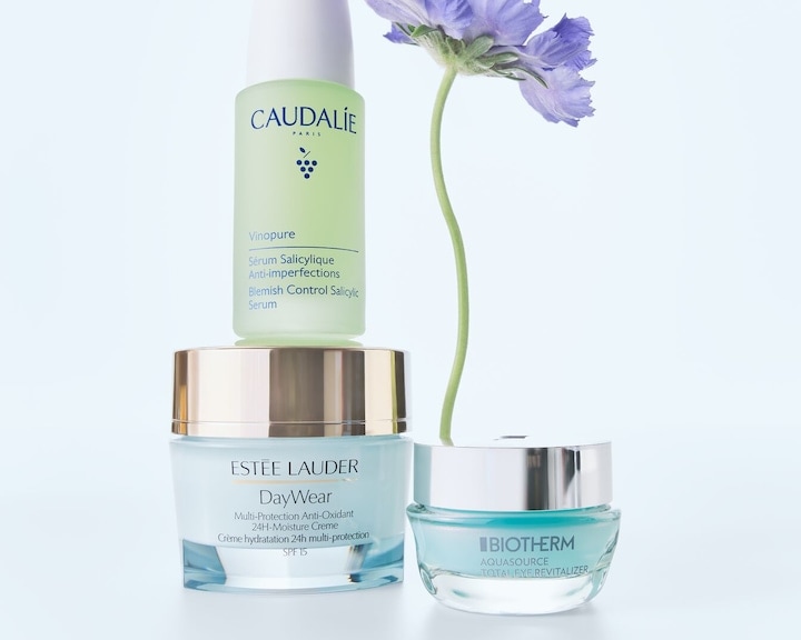 Produse esențiale pentru îngrijirea pielii, incluzând serul Caudalie Vinopure, crema hidratantă Estée Lauder DayWear și crema Biotherm Aquasource, evidențiate de o floare mov.