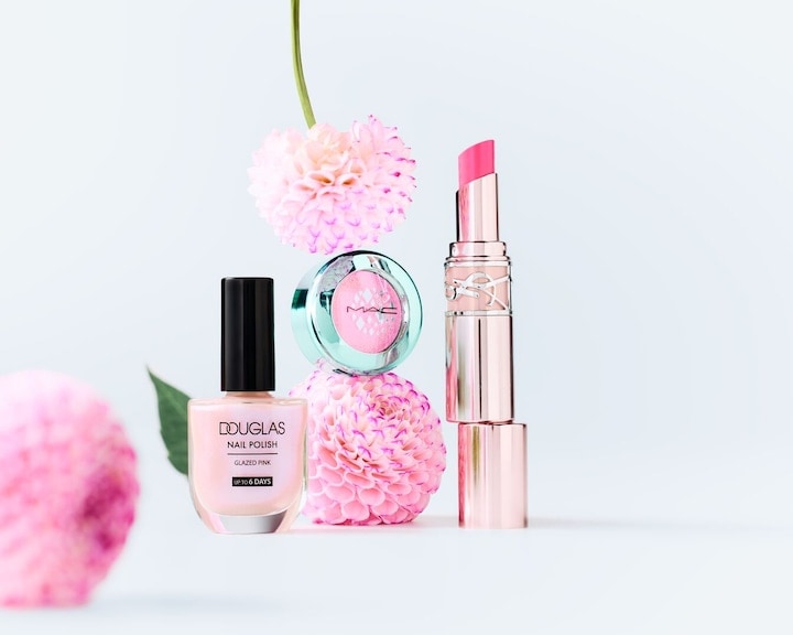 O colecție de produse de beauty în nuanțe de roz, inclusiv ojă Douglas în roz glazurat, fard de pleoape MAC Cosmetics și ruj Yves Saint Laurent, sunt aranjate alături de flori roz.